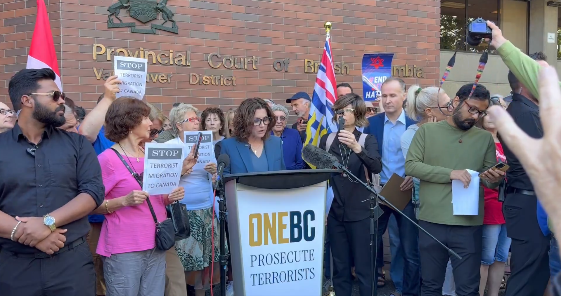 Vancouver MLA Dallas Broide speaking at a press conference outside Vancouver Provincial Court, making a public announcement today about taking action against Charlotte Kates and Samidoun. Broide is standing at a podium, addressing media and supporters, with the courthouse visible in the background.